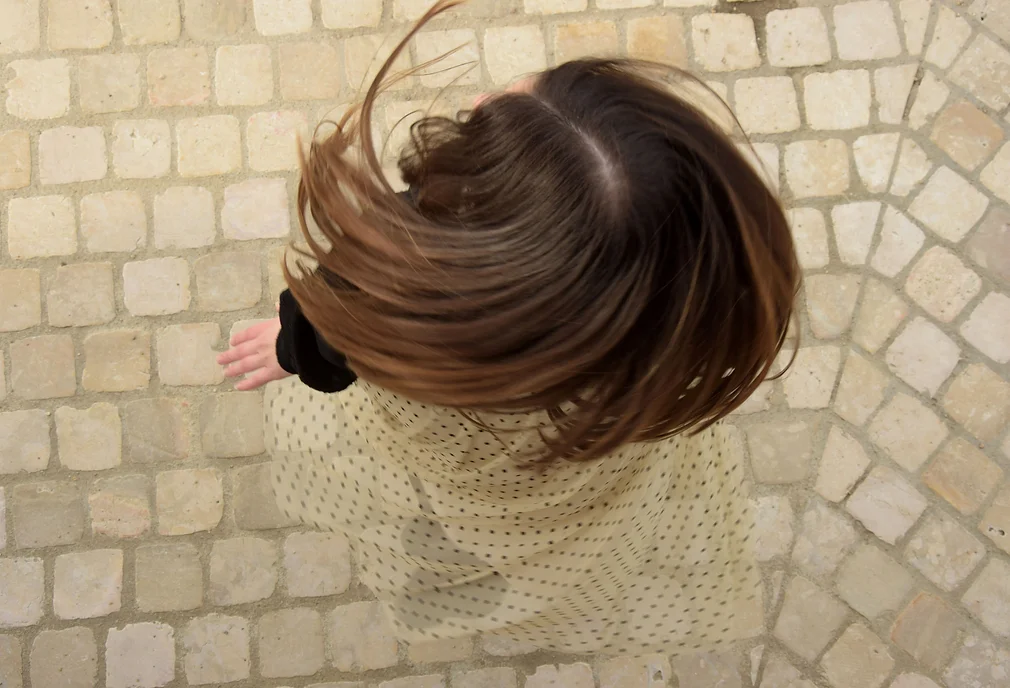 Photo prise d'en haut d'une femme brune qui tourne sur elle même faisant tourner ses cheveux et sa jupe beige à pois noir. Au sol des pavés de couleur beige.