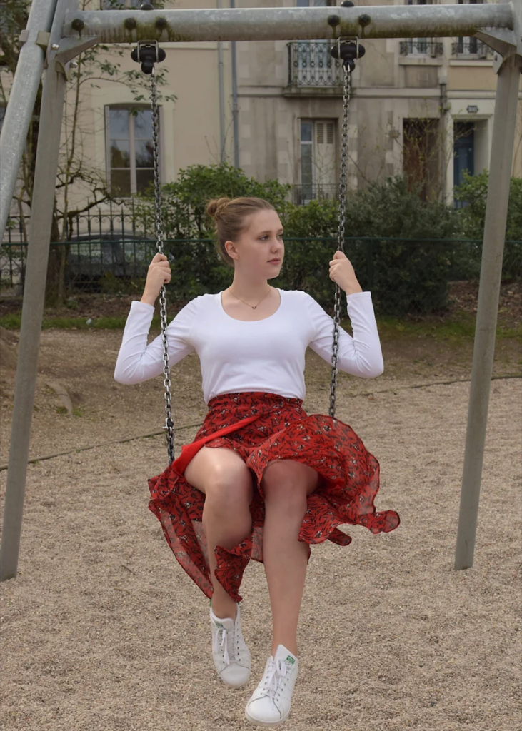 Photo d'une jeune femme blonde sur une balançoire. Haut blanc, jupe rouge qui vole grâce au mouvement de la balançoire.