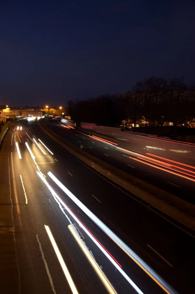 Photographie prise d'un pont qui surplombe une route. Les lumières des voitures circulant forment des faisceaux lumineux. La photo est divisée en deux à droite une route avec des lumières rouges à gauche des lumières blanches.