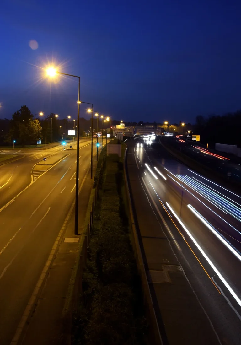 Photographie prise d'un pont qui surplombe une route. Les lumières des voitures circulant forment des faisceaux lumineux. La photo est divisée en deux à droite une route avec de la circulation plongée dans le noir à gauche aucune voiture et la route est éclairée.